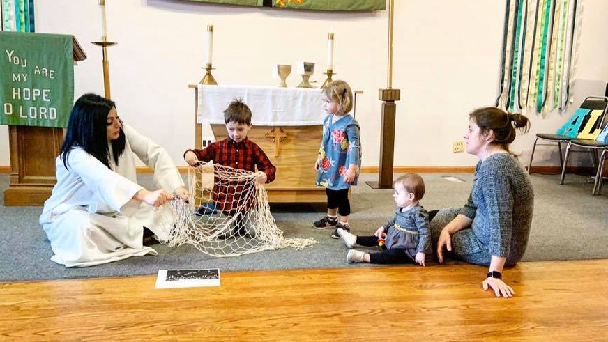 Pastor Elle and a child with short, brown hair hold up a net during a children's sermon. Two children and a parent watch.